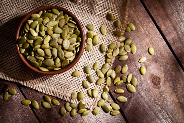 Top view of a brown bowl filled with organic pumpkin seeds shot on rustic wood table. Some almonds are out of the bowl on a burlap. Predominant colors are green and brown. DSRL studio photo taken with Canon EOS 5D Mk II and Canon EF 100mm f/2.8L Macro IS USM